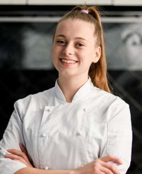 A young woman in a white chef's uniform standing in front of an oven.