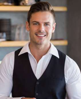 A smiling man in a vest standing in front of a bar.